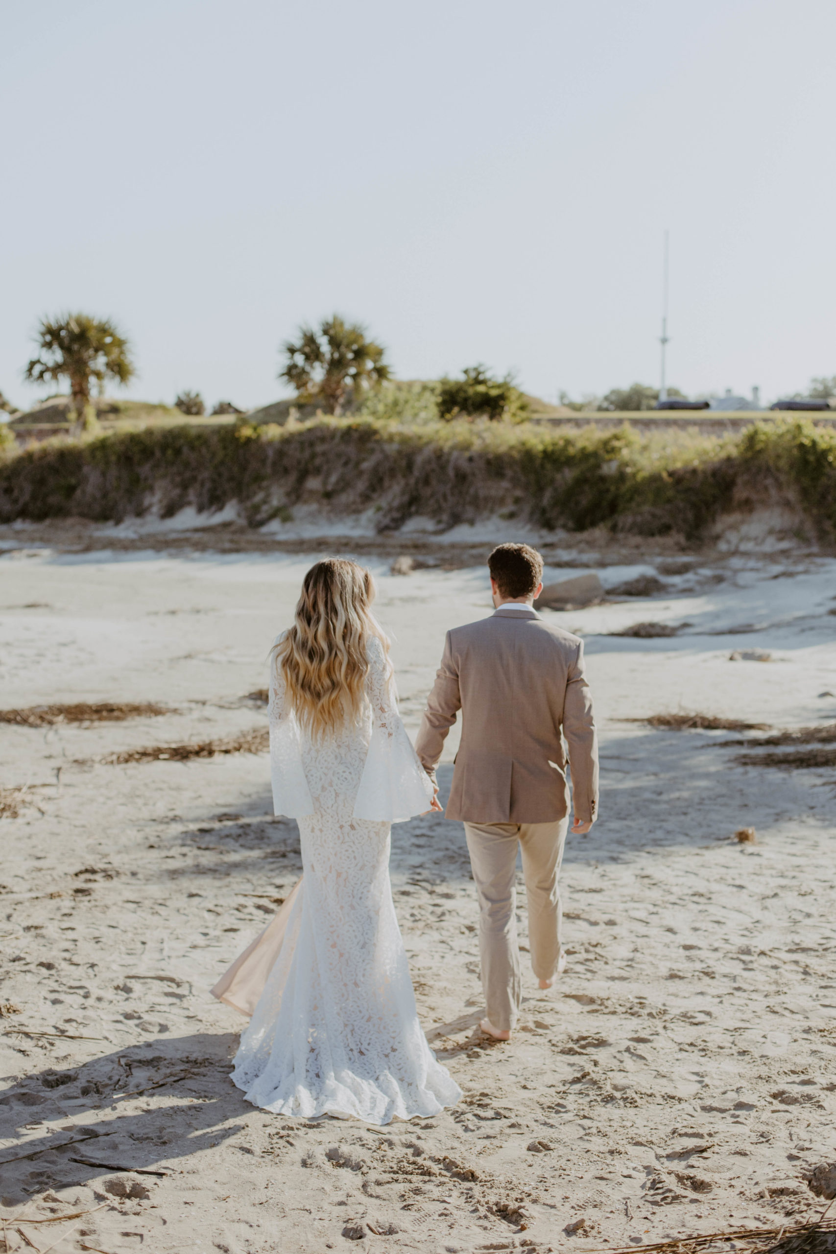 Bride wearing beautiful bohemian dress and groom wearing neutral tan suit walking along the beach barefoot away from the camera while holding hands.