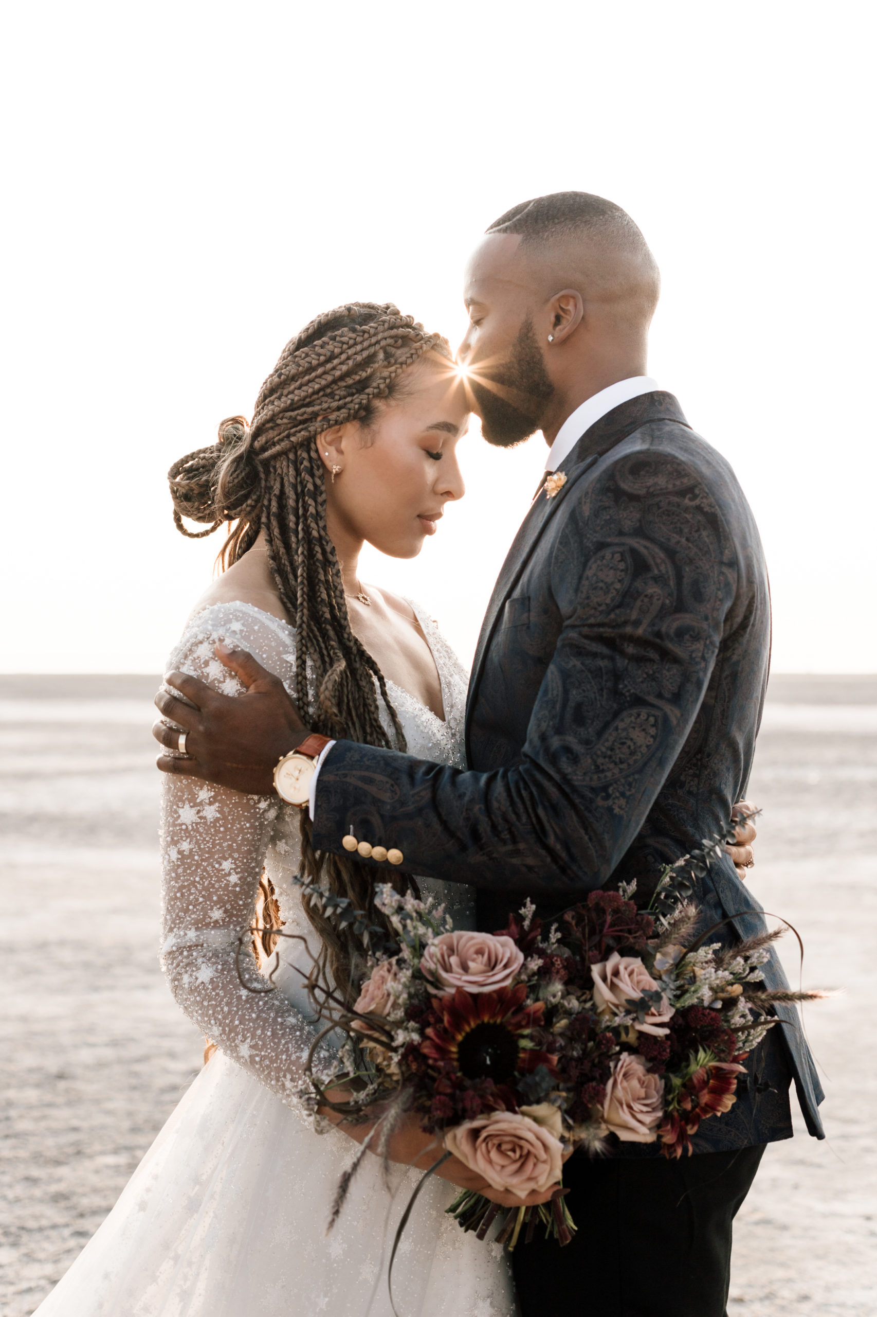 Groom wearing velvet suit kissing bride wearing boho mesh wedding dress on her forehead during their golden hour elopement at the Bonneville Salt Flats in Utah.