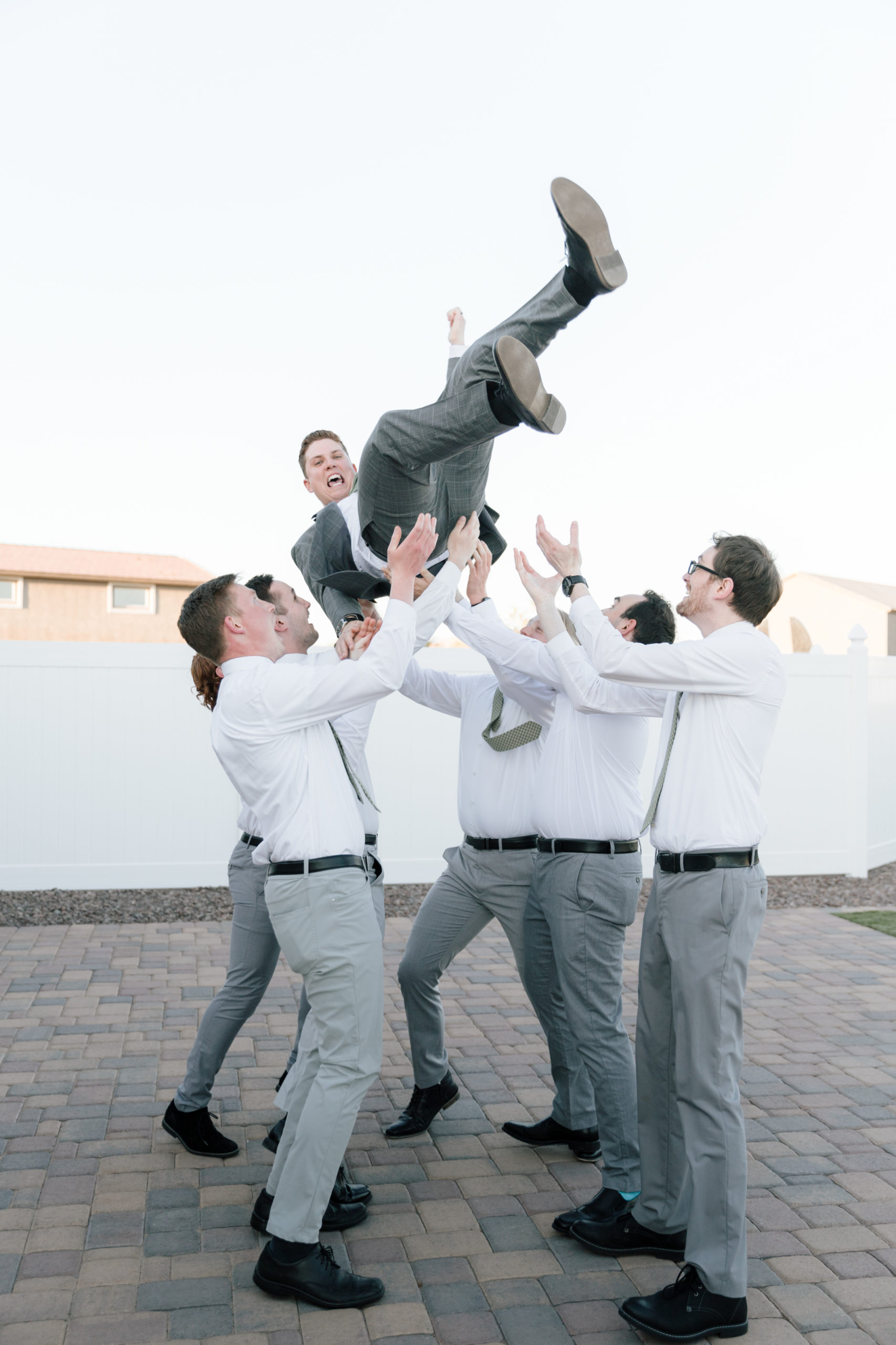 Groomsmen wearing gray pants and white button down shirts throwing groom in the air celebrating during backyard wedding.