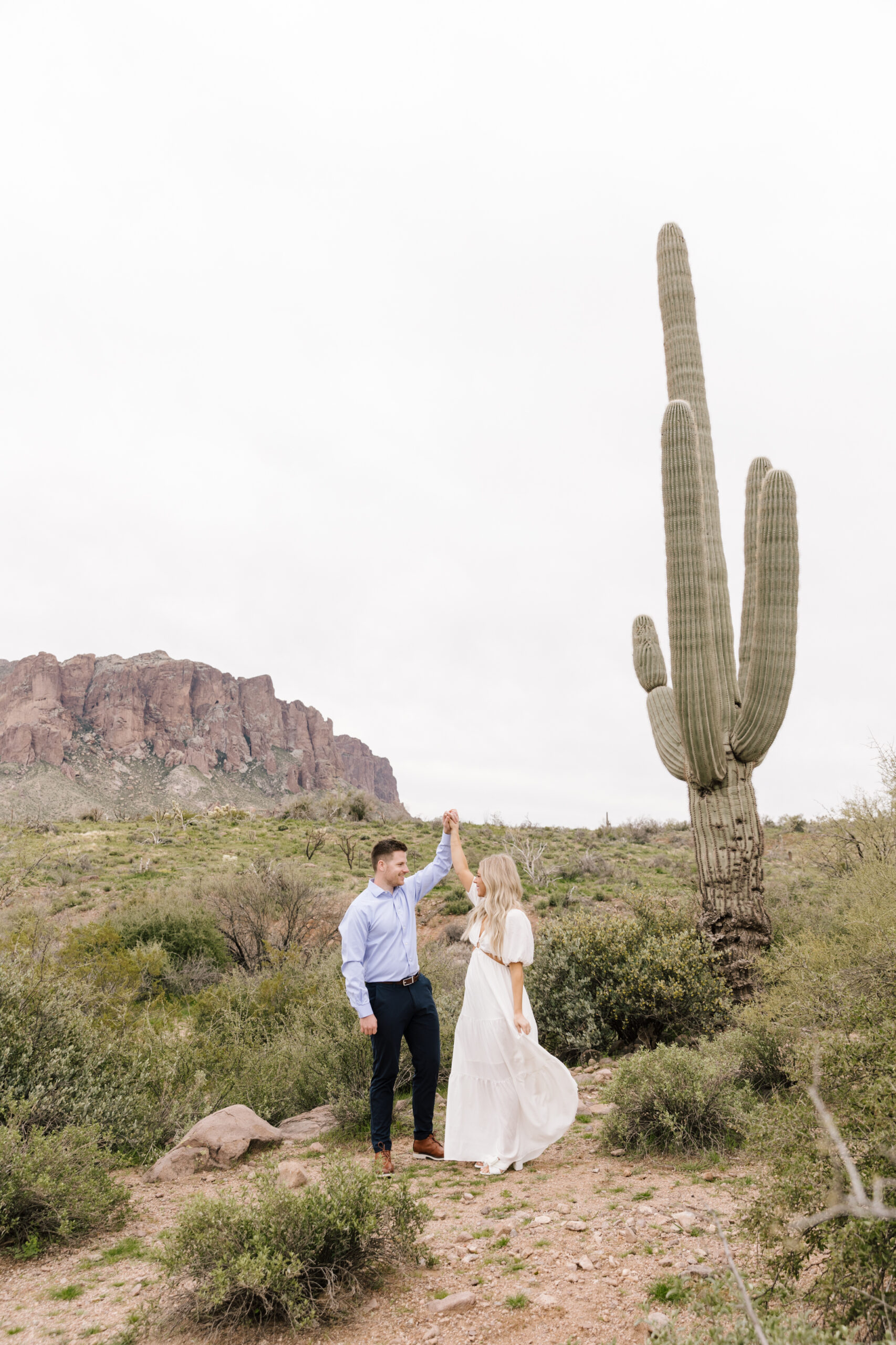 Guy twirling is fiance wearing a white dress in front of a tall Saguaro cactus at Lost Dutchman State Parl