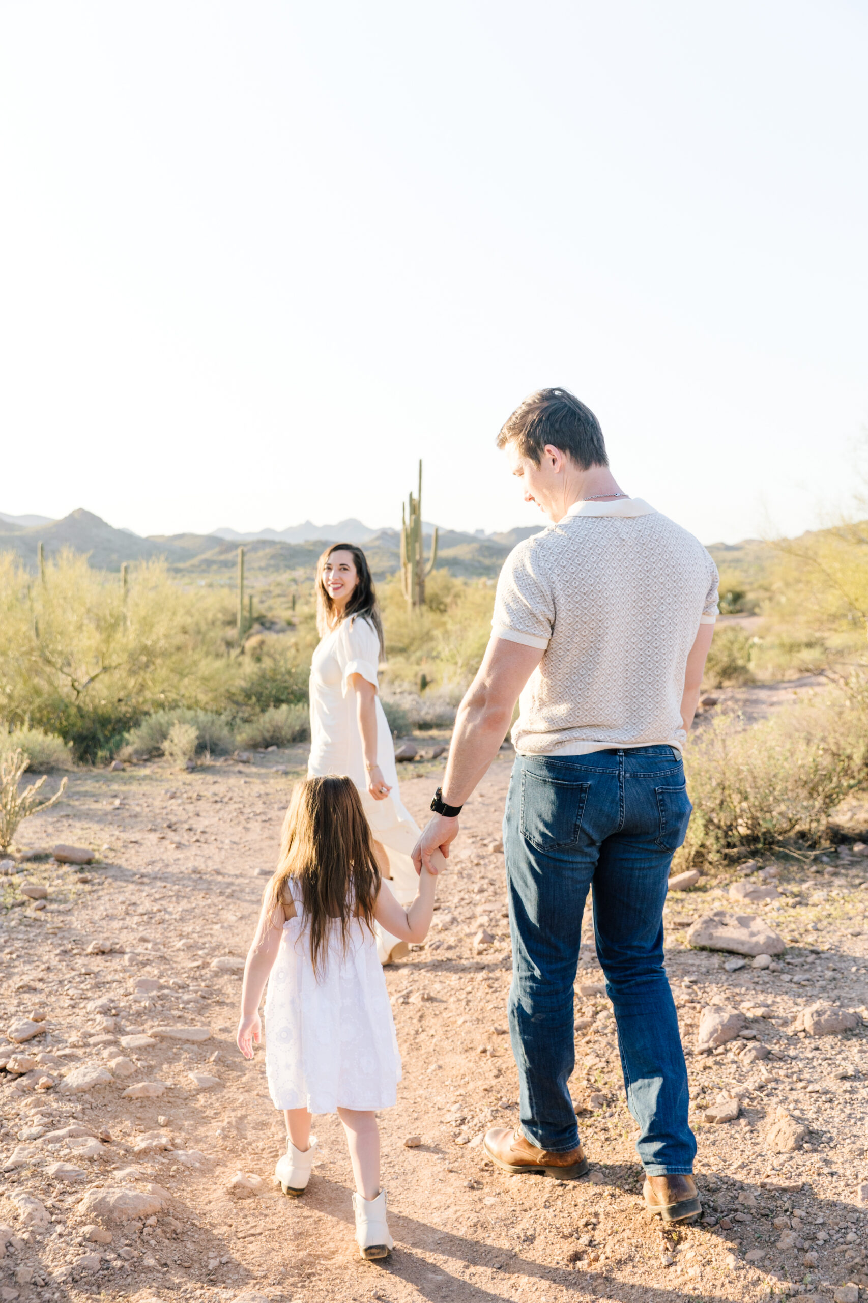 Family of 3 smiling and walking together down a desert path with the mountains and cacti in the background at Lost Dutchman State Park
